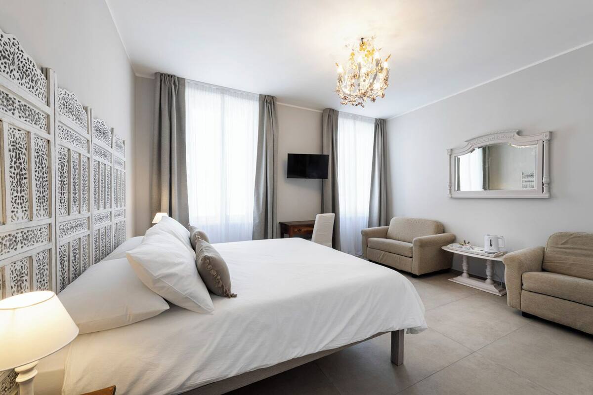 A hotel room with a large bed, a desk, two armchairs, a TV, and a chandelier.