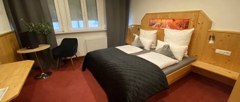 A compact hotel room with a bed, a small desk, a chair, a potted plant, and a window with curtains.