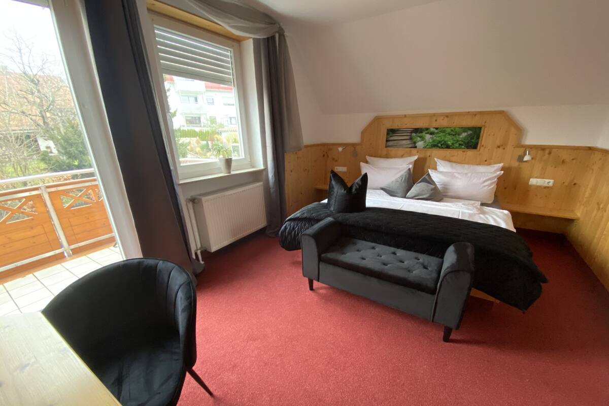 A bedroom with a bed, a black armchair, a wooden headboard, a window with blinds, and a red carpet.