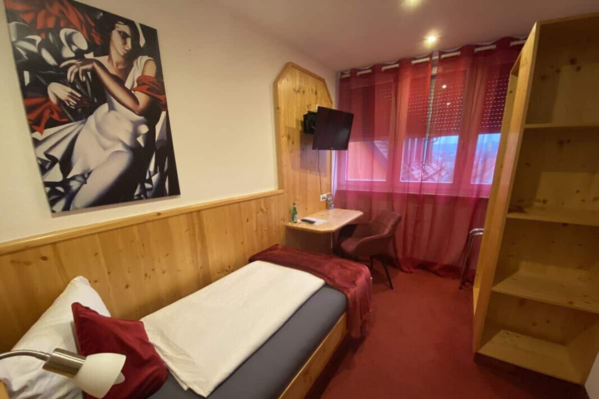 A single bed with a red blanket, a desk with a chair, a TV mounted on the wall, and a wooden bookshelf.