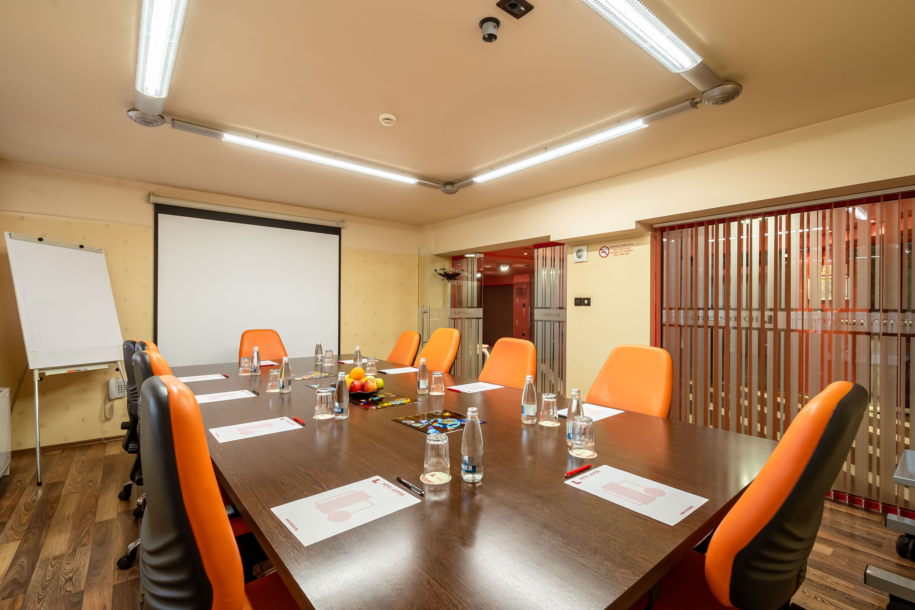 Une salle de conférence avec une longue table, des chaises orange, un tableau blanc et un écran de projection.