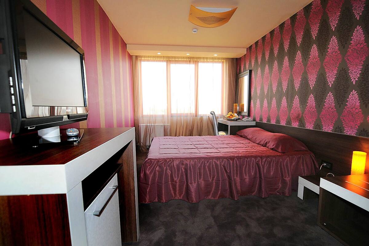 Quarto de hotel com cama, escrivaninha com computador, televisão e janela com cortinas.