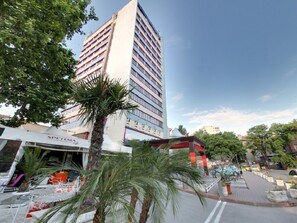 Un hotel alto de diseño moderno, con una entrada destacada y un restaurante visible llamado "SPETEMA" en la planta baja.