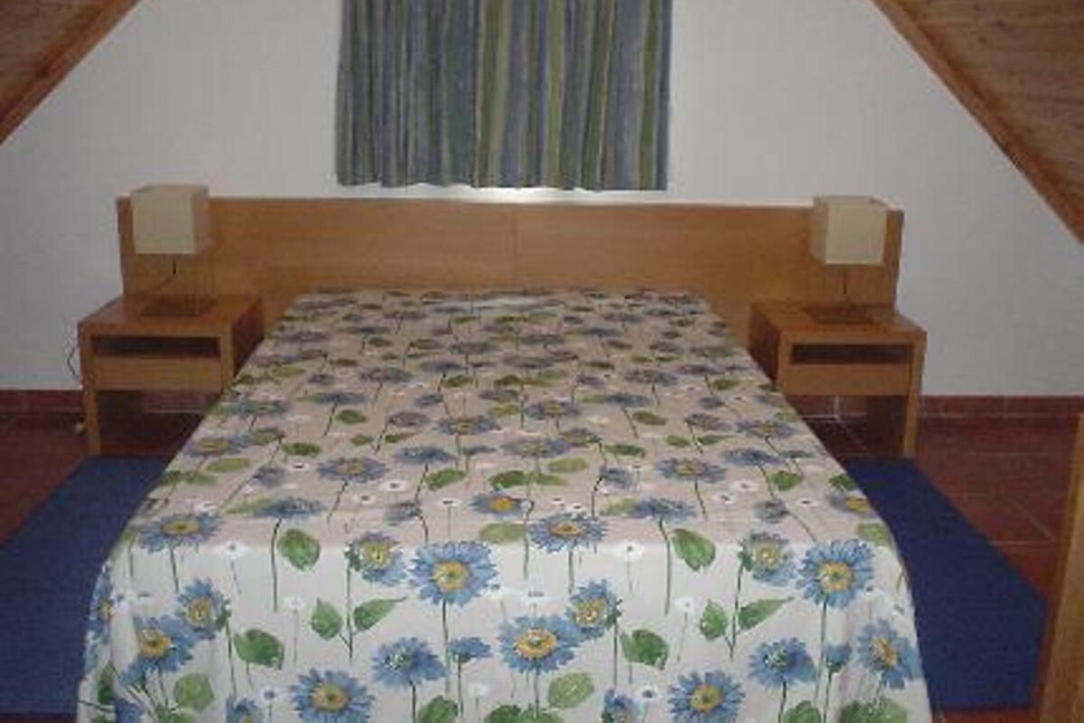A bedroom with a wooden headboard, a floral bedspread, two bedside tables with lamps, and a window with curtains.