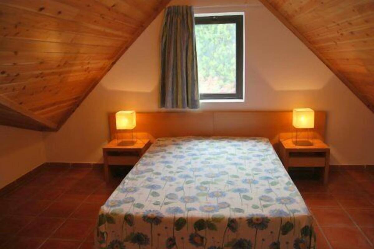 A small, cozy attic bedroom with a sloped ceiling, a single bed with a floral bedspread, two bedside lamps, and a window with curtains.