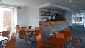 A modern dining area with orange chairs and tables, a central counter with a coffee machine, and a view of the outdoors. - Apartamentos Turísticos Nossa Senhora da Estrela (Lagoa)