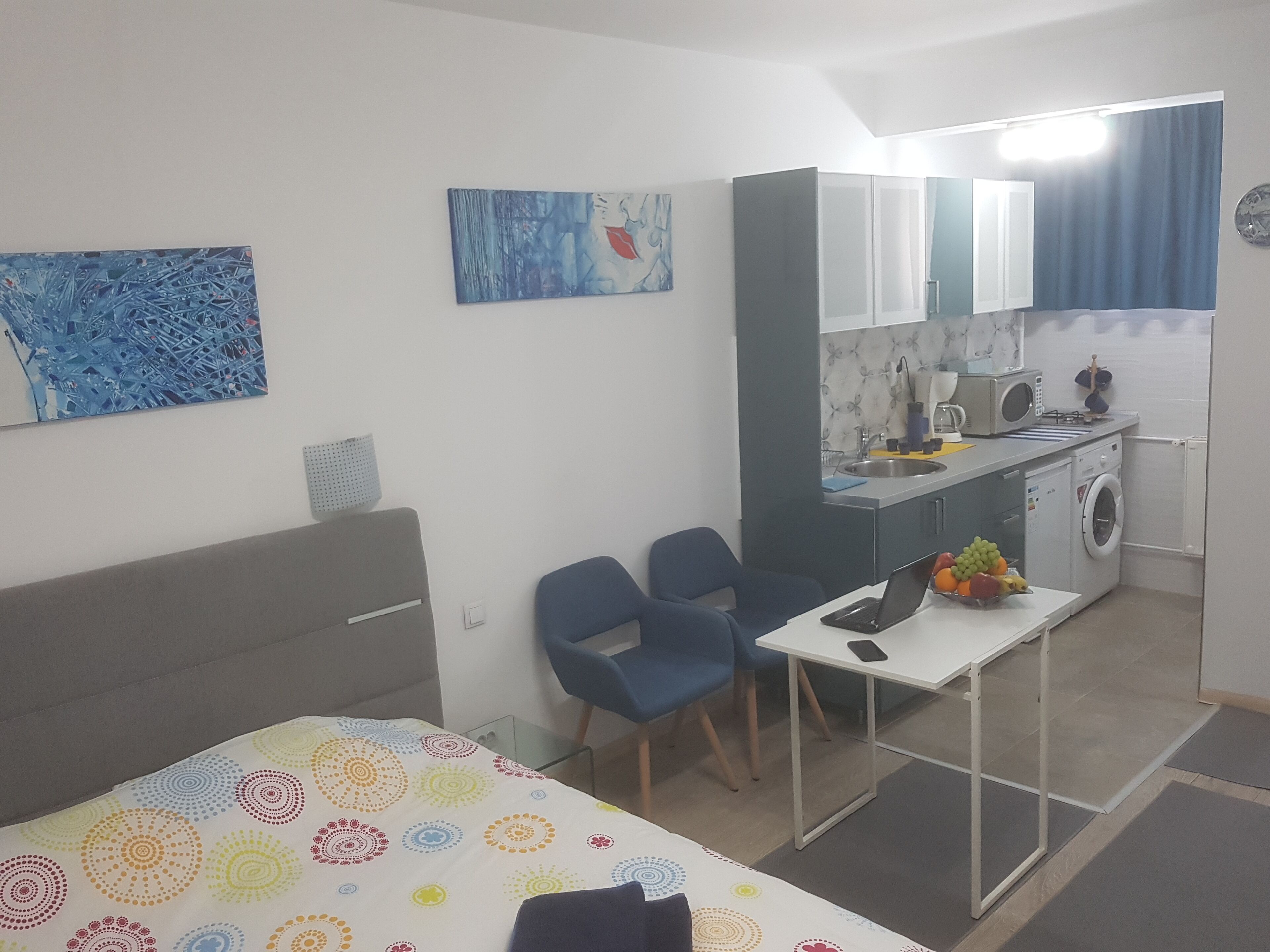 Ein kompaktes Hotelzimmer mit Bett, kleinem Schreibtisch, Stuhl, Küchenzeile und Waschmaschine.