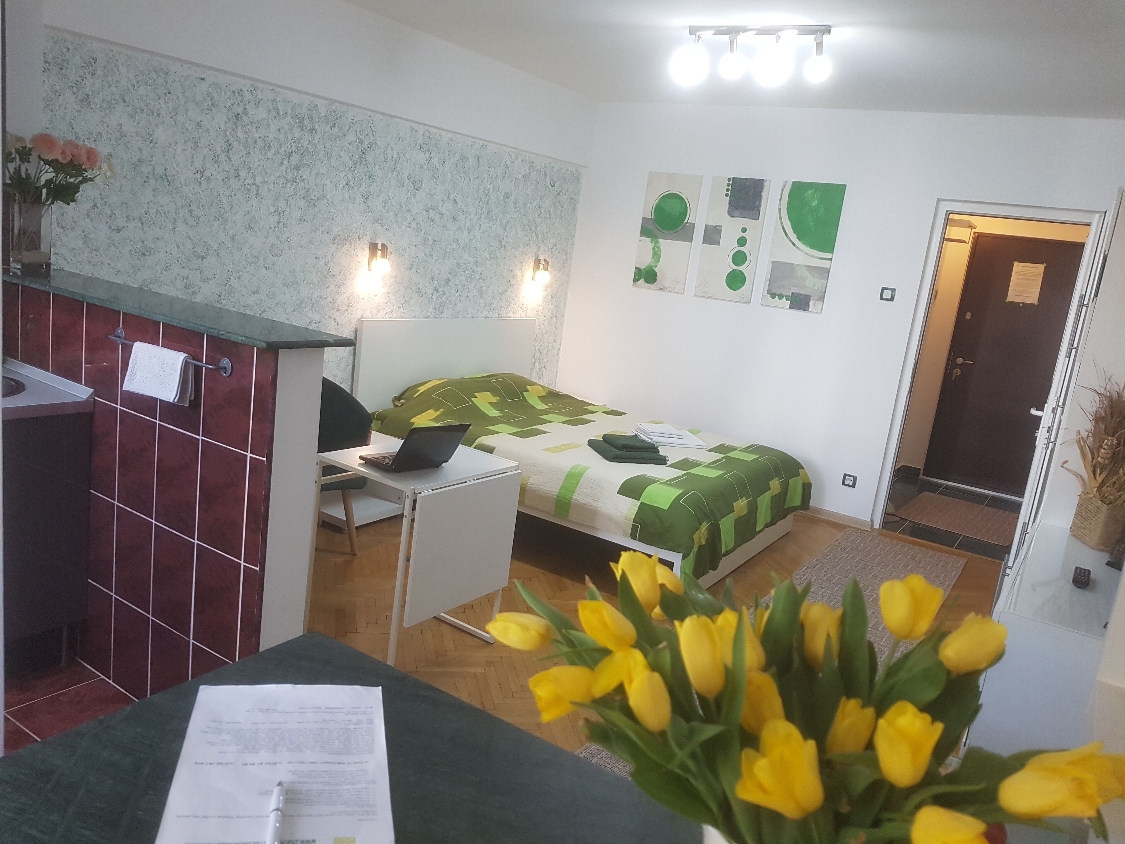 Una stanza con un letto, una scrivania, una sedia, un tavolino con un vaso di fiori gialli e una porta.