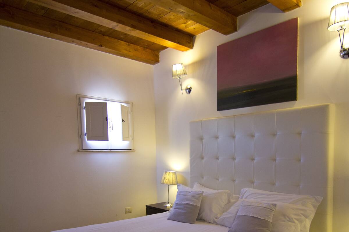 Une chambre dotée d’un grand lit, de poutres apparentes en bois au plafond, d’une fenêtre offrant une belle vue et d’un luminaire fixé au mur.