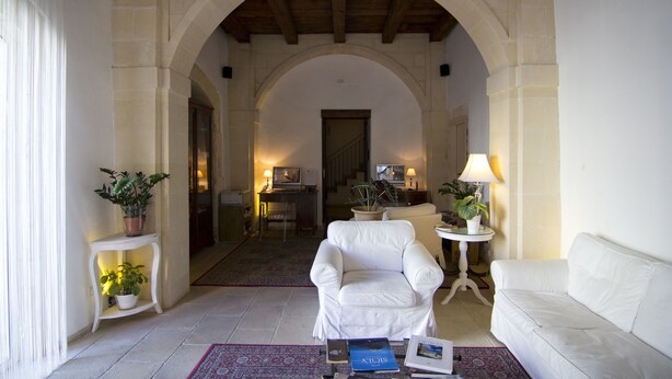 A spacious living area with a white sofa, a white armchair, and a small table with books. There are potted plants on side tables and a lamp on a side table. An arched doorway leads to another room. - B&B La Via della Giudecca (Syracuse)
