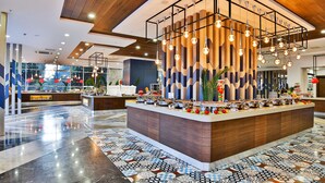 Hall d'hotel moderna con un'area buffet centrale, un'ampia finestra vista su vegetazione e un pavimento decorato.