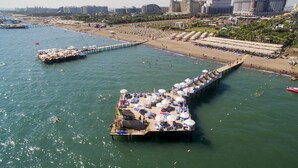 Un molo con ombrelloni e sdraio che si estende su una spiaggia, con uno skyline urbano sullo sfondo.