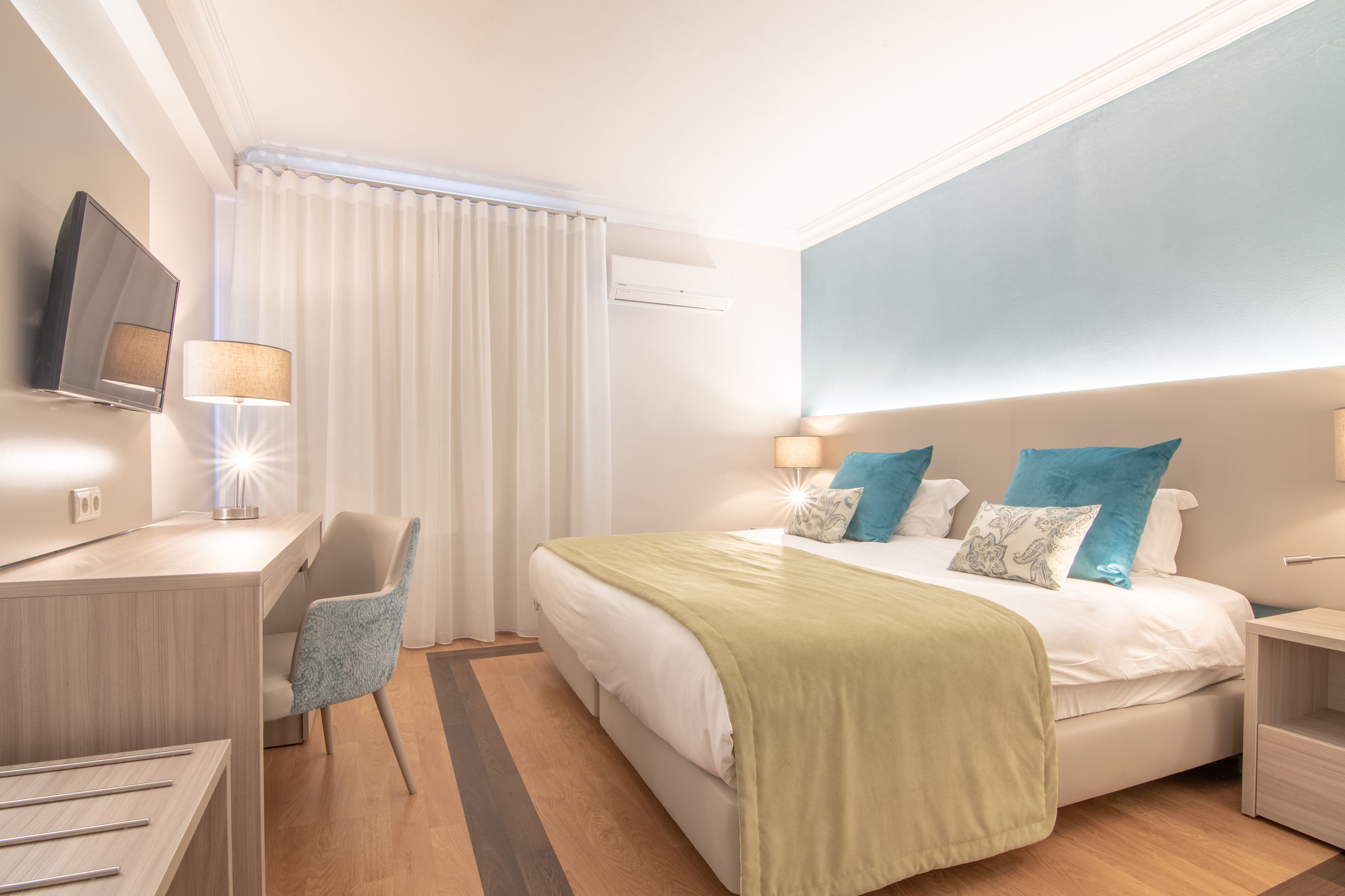 A modern hotel room with a bed, bedside tables, a desk, a chair, a TV, and a window with curtains.