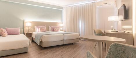 Habitación de hotel con cama doble, cama individual, escritorio con silla y televisión montada en la pared.