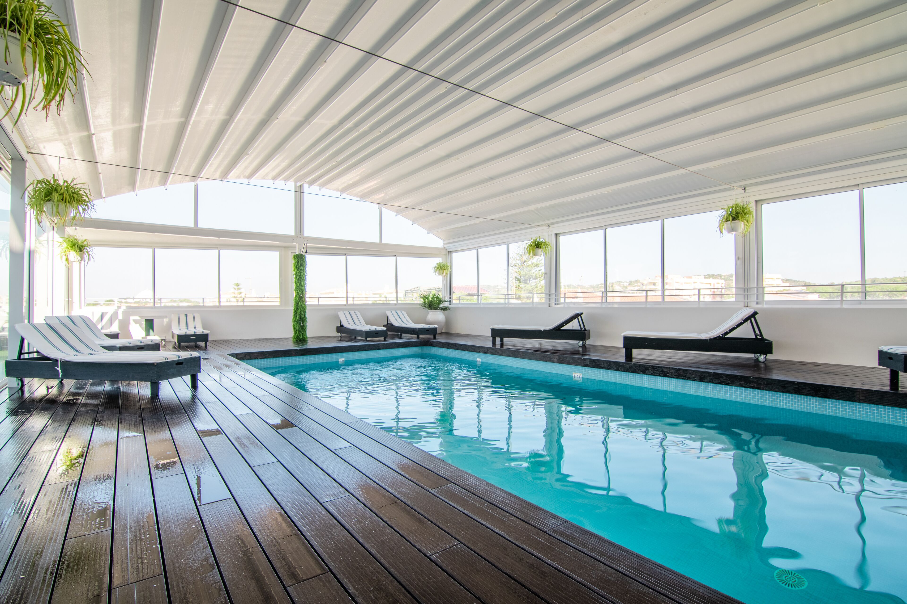 An indoor swimming pool with wooden decking, lounge chairs, and potted plants.