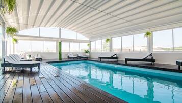 An indoor swimming pool with wooden decking, lounge chairs, and potted plants.