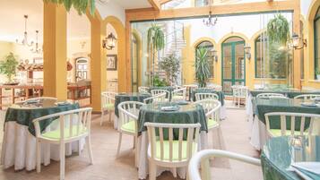 Een restaurant met ronde tafels en witte stoelen, groene tafelkleden en hangende planten.