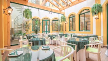 Un restaurante con mesas y sillas redondas, manteles verdes, plantas colgantes y ventanales arqueados.
