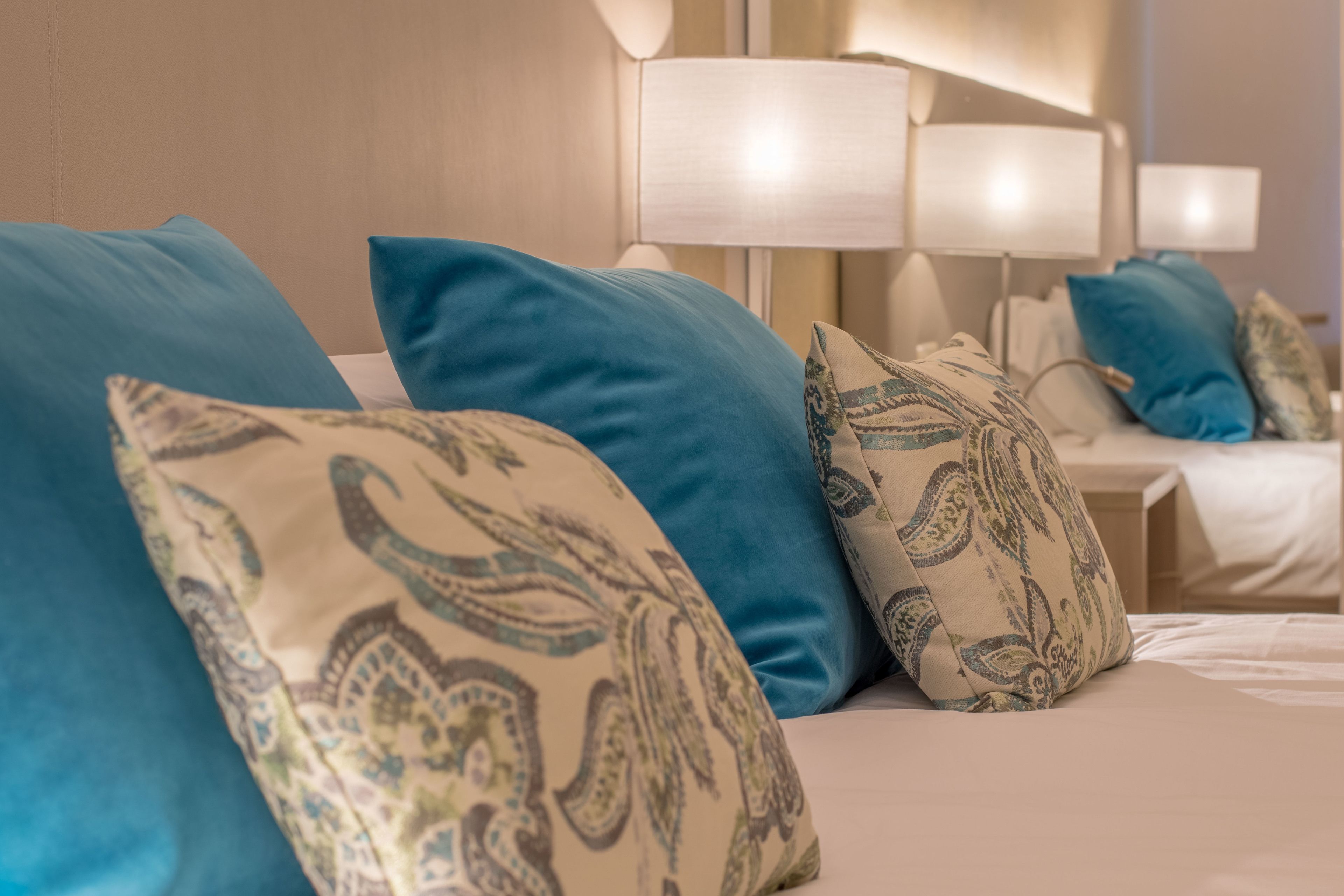 A hotel room with a bed featuring blue and patterned pillows, a white headboard, and a bedside lamp.