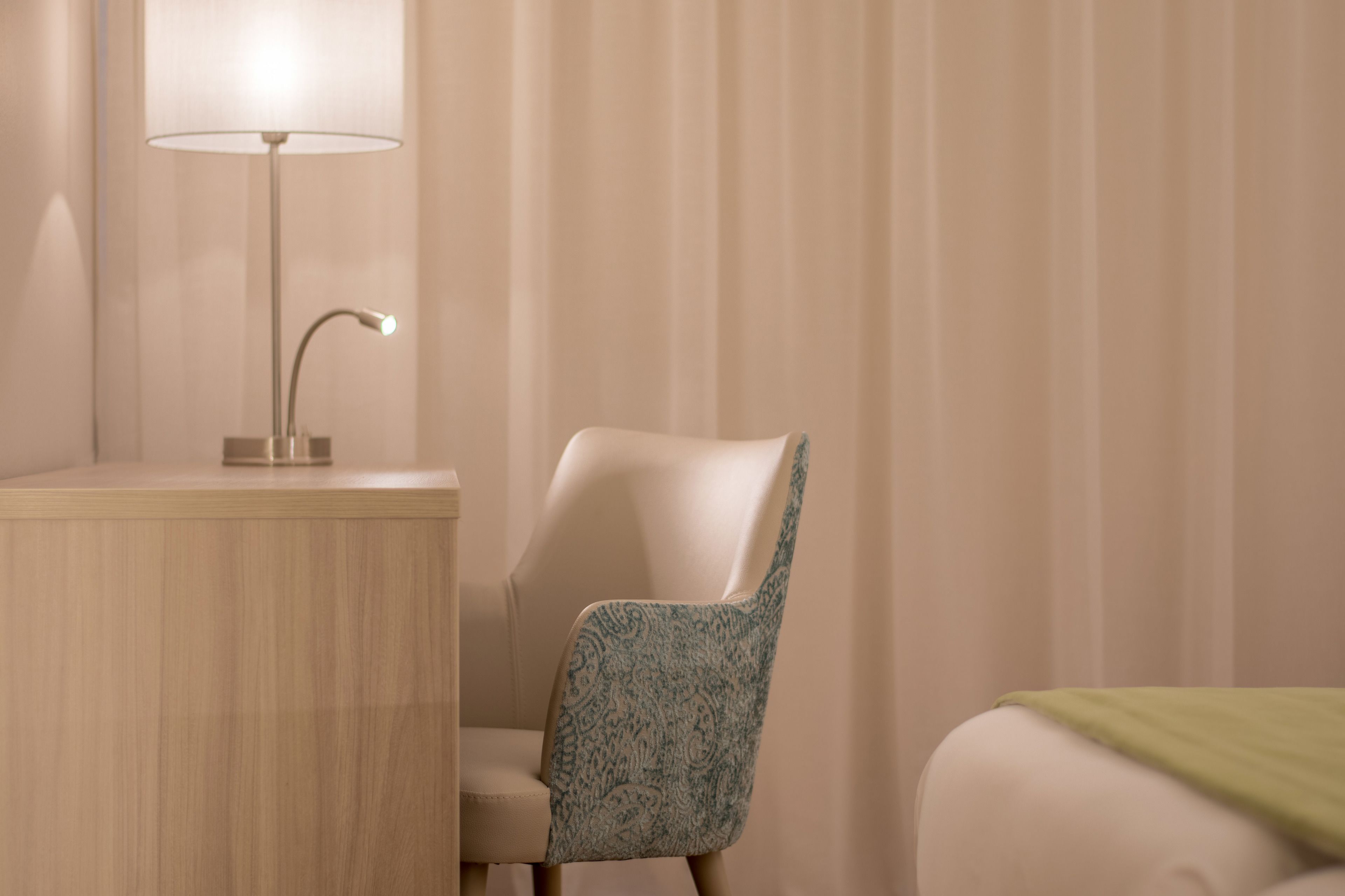A hotel room with a desk, a chair with a patterned cushion, a lamp, and a bed with a green blanket.