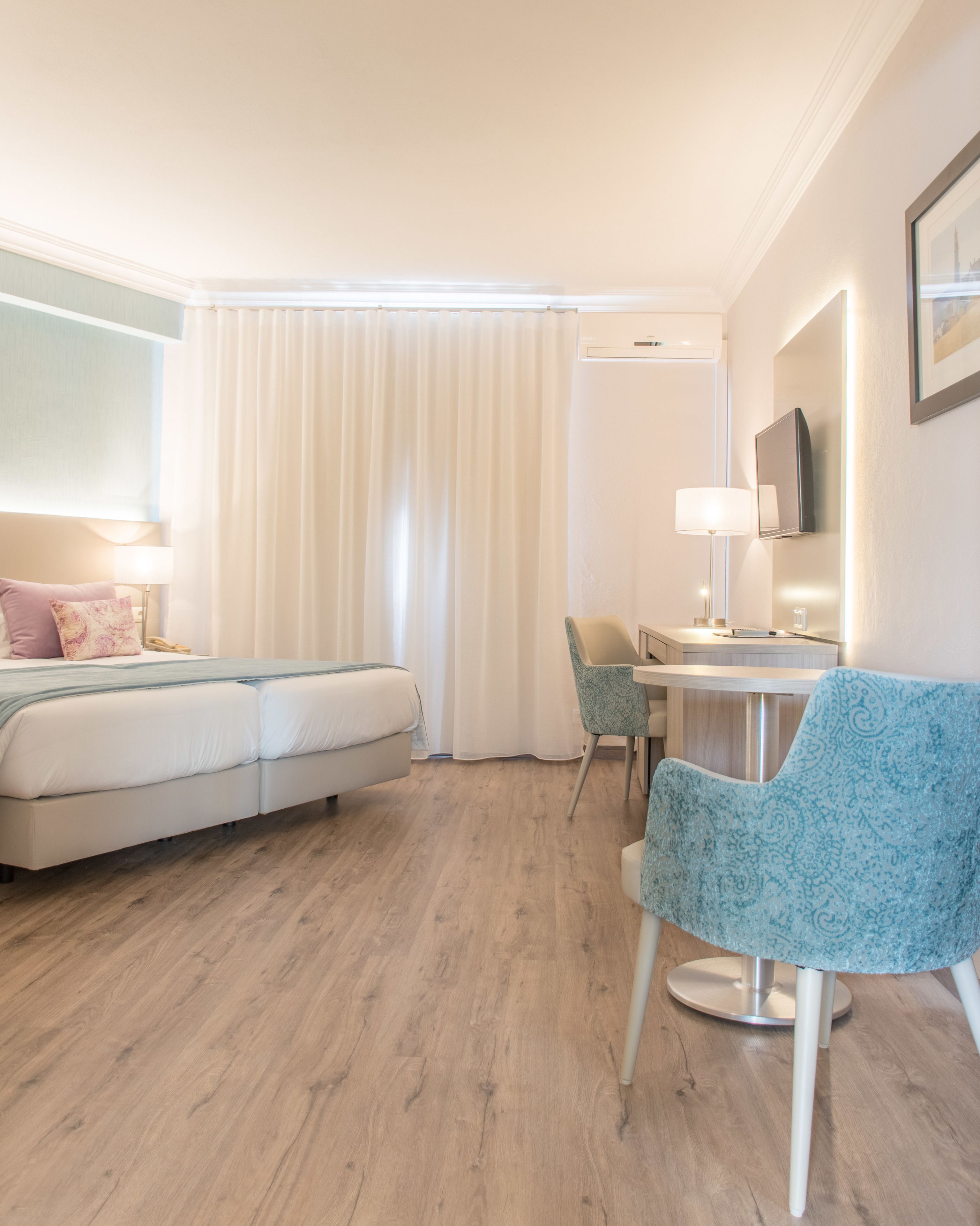 A hotel room with a bed, a desk with a chair, a TV, and a window with curtains.