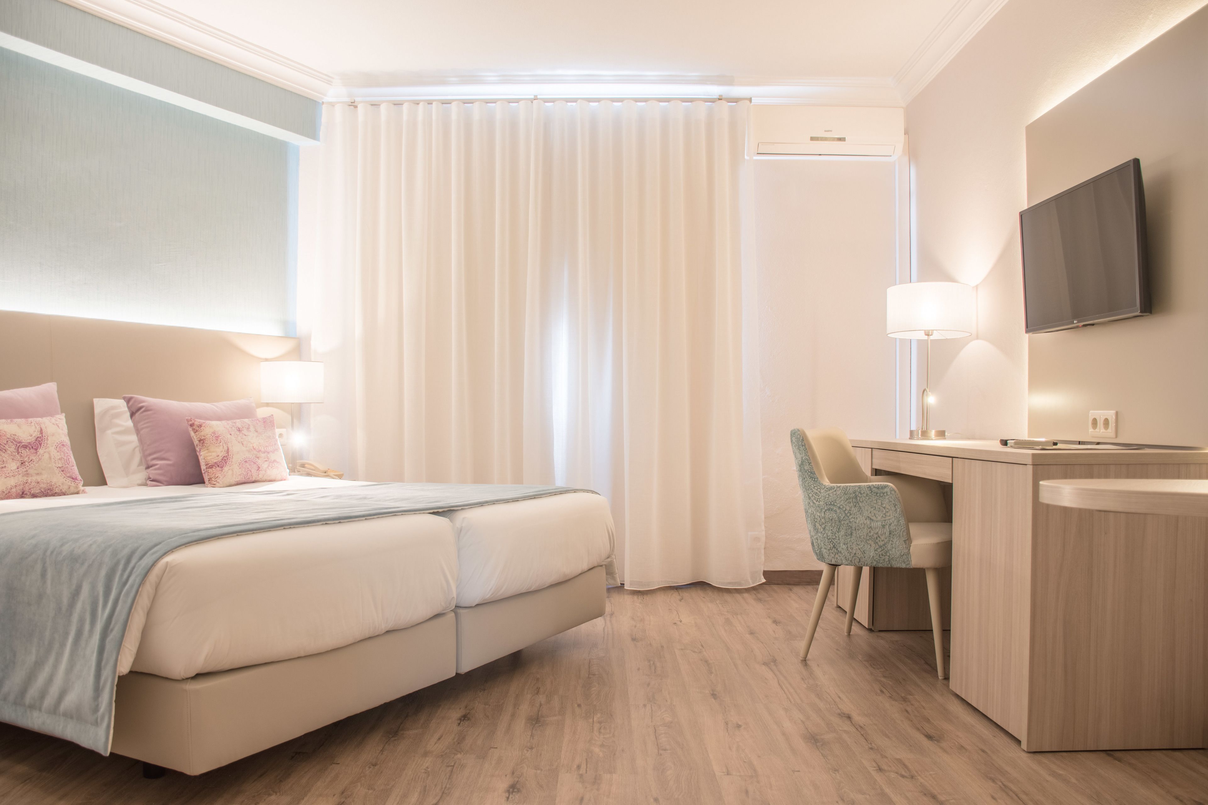 A hotel room with a bed, a desk, a chair, a TV, and a window with curtains.