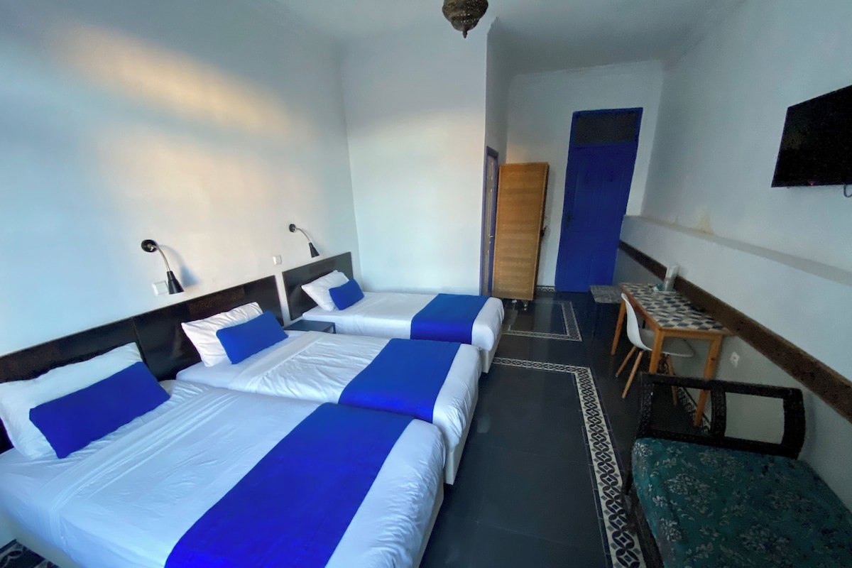A hotel room with three beds, a small table, and a bench.