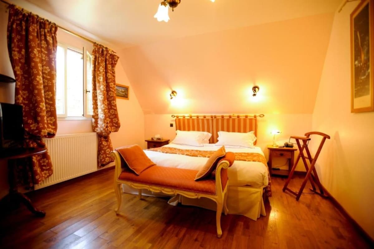 Une chambre à coucher avec un lit double, une table de chevet, une chaise, un radiateur et une fenêtre avec des rideaux.