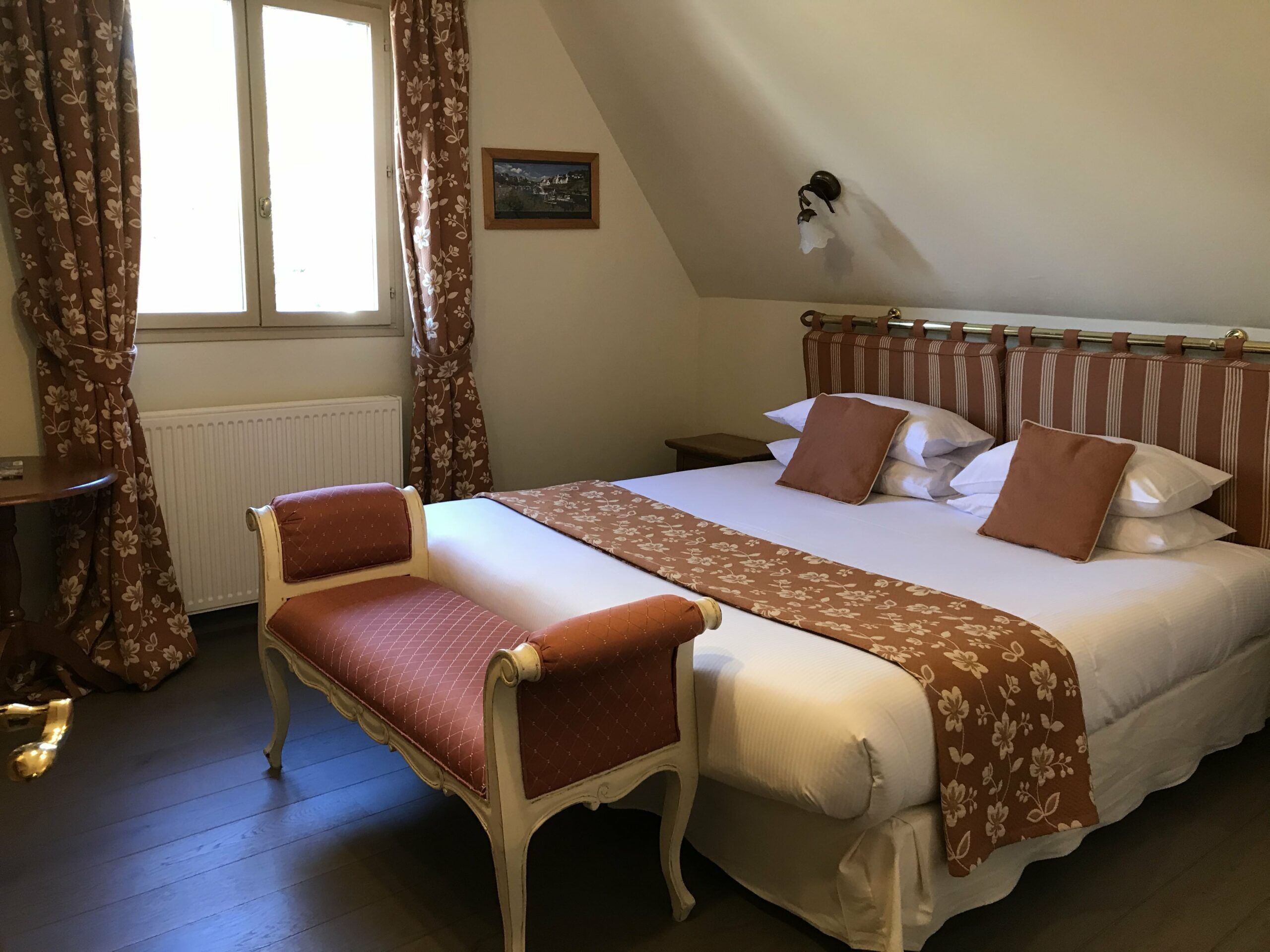 Un dormitorio con cama, un banco, un radiador, una ventana y cortinas.