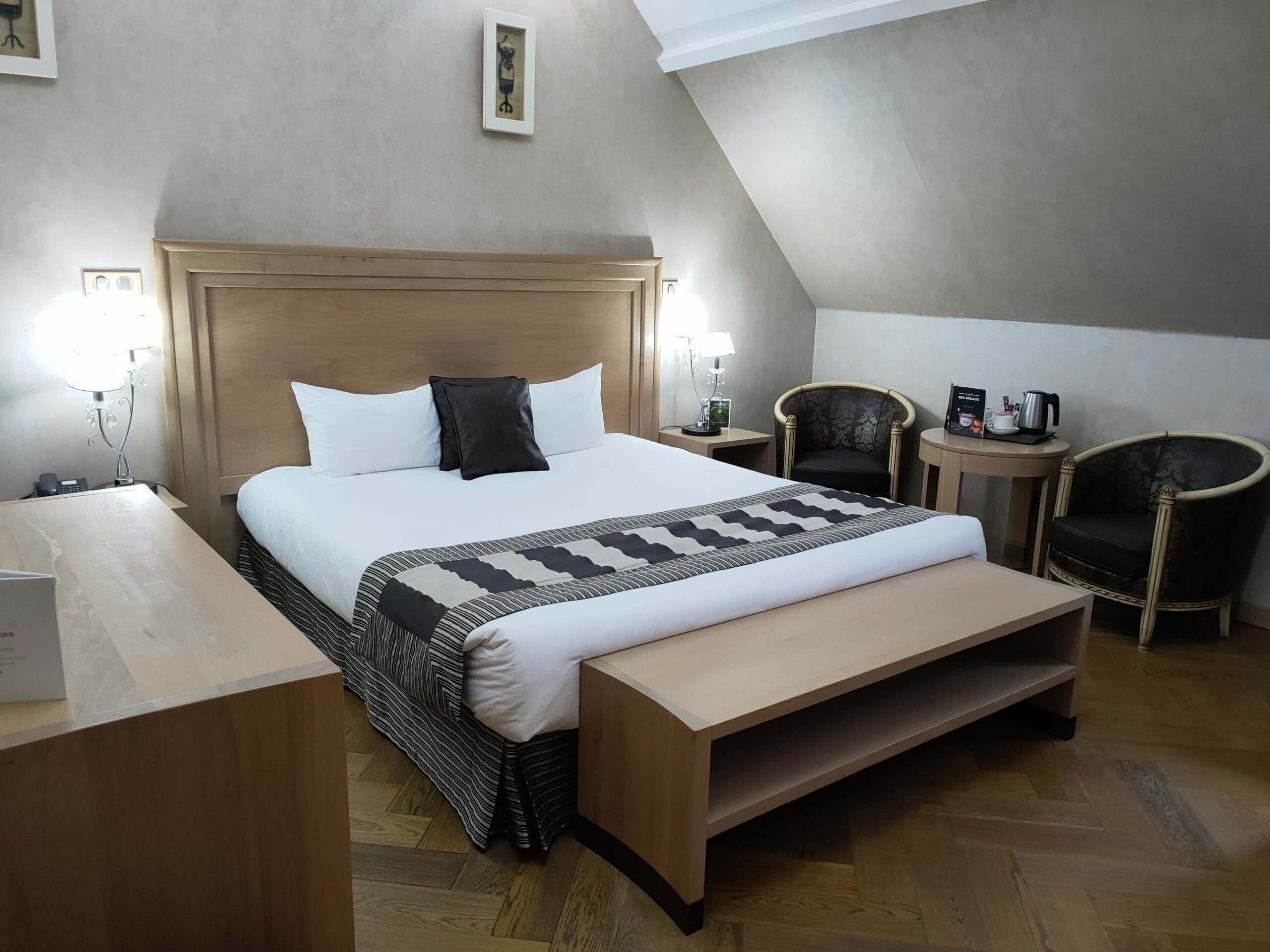 A hotel room with a large bed, a wooden bench, two chairs, a small table with a kettle, and a framed picture on the wall.
