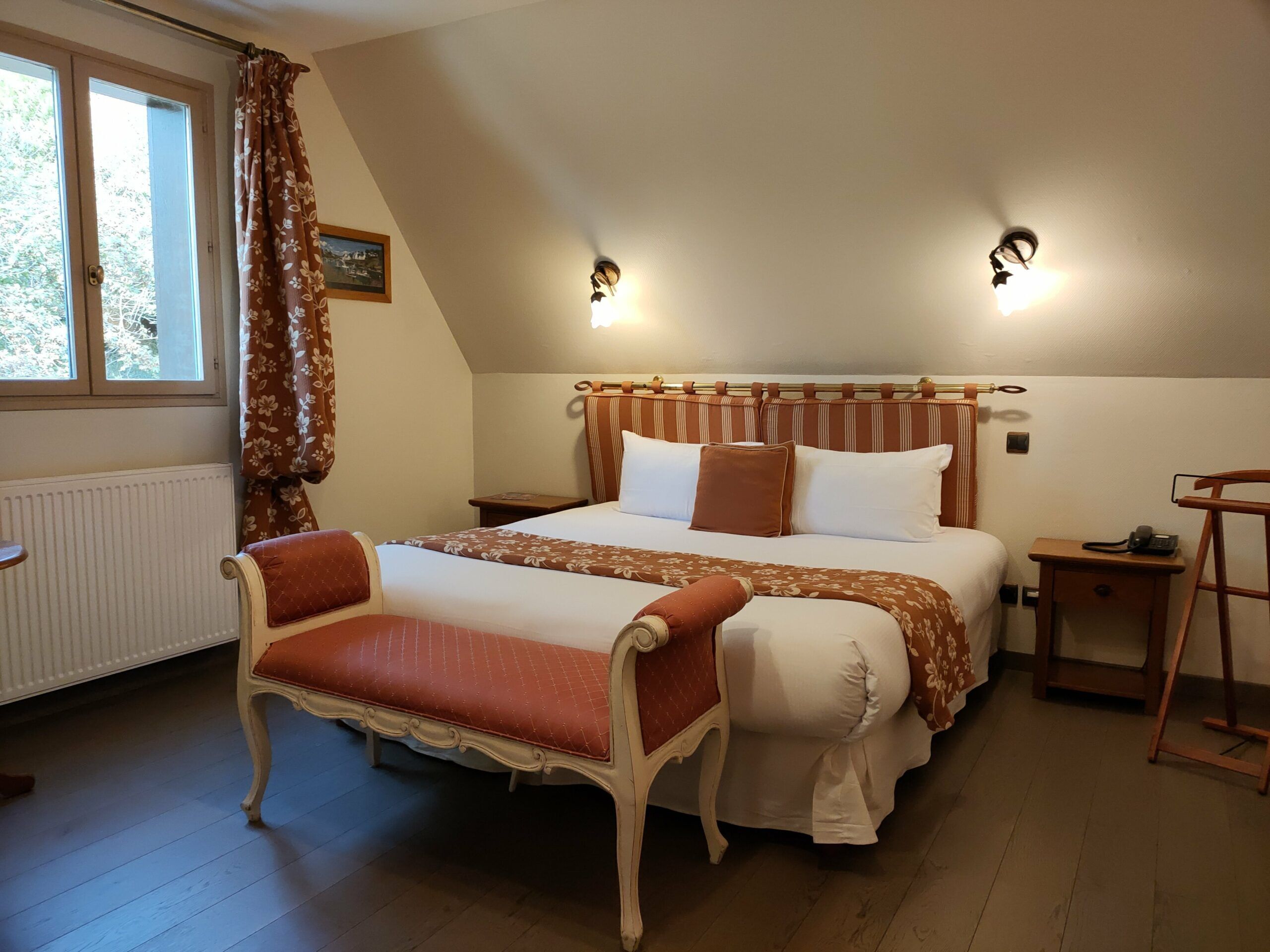Ein Schlafzimmer mit einem Bett, einer Bank, einem Nachttisch, einer Lampe und einem Fenster mit Vorhängen.