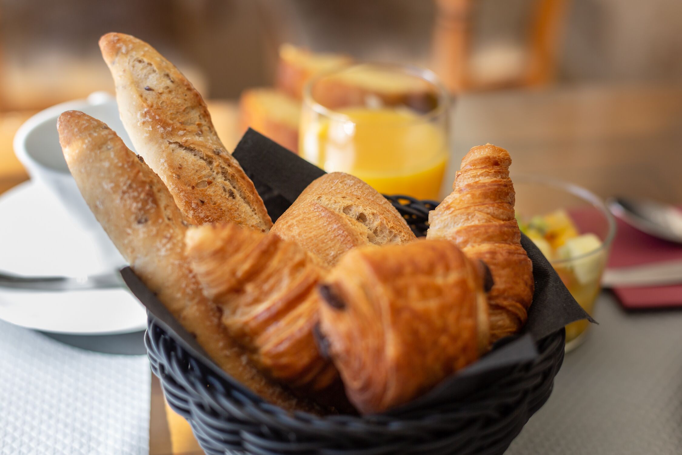 A basket of croissants and breadsticks on a table with a glass of orange juice and a plate.