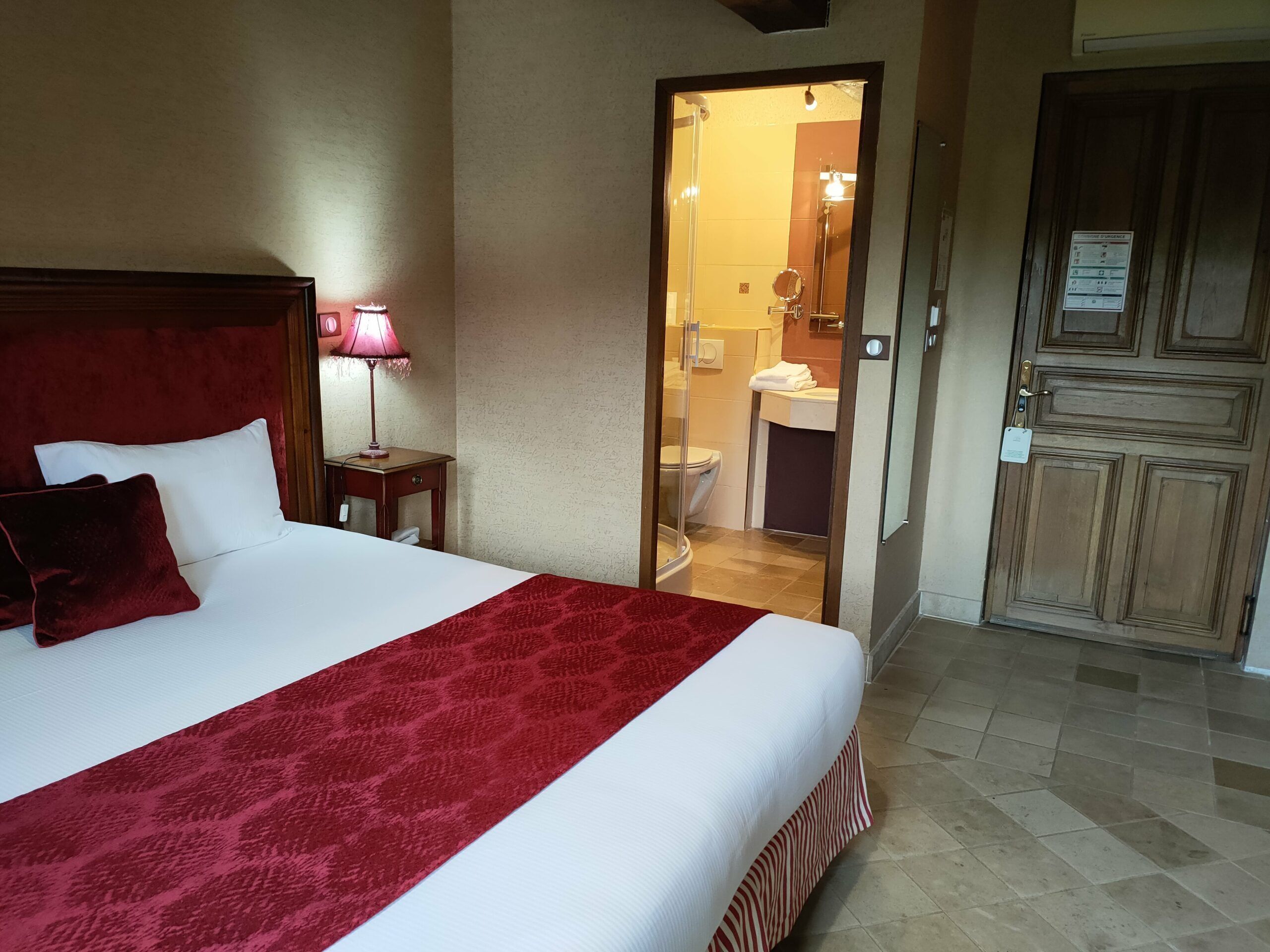 A hotel room with a large bed, a bedside table with a lamp, and a door leading to a bathroom.