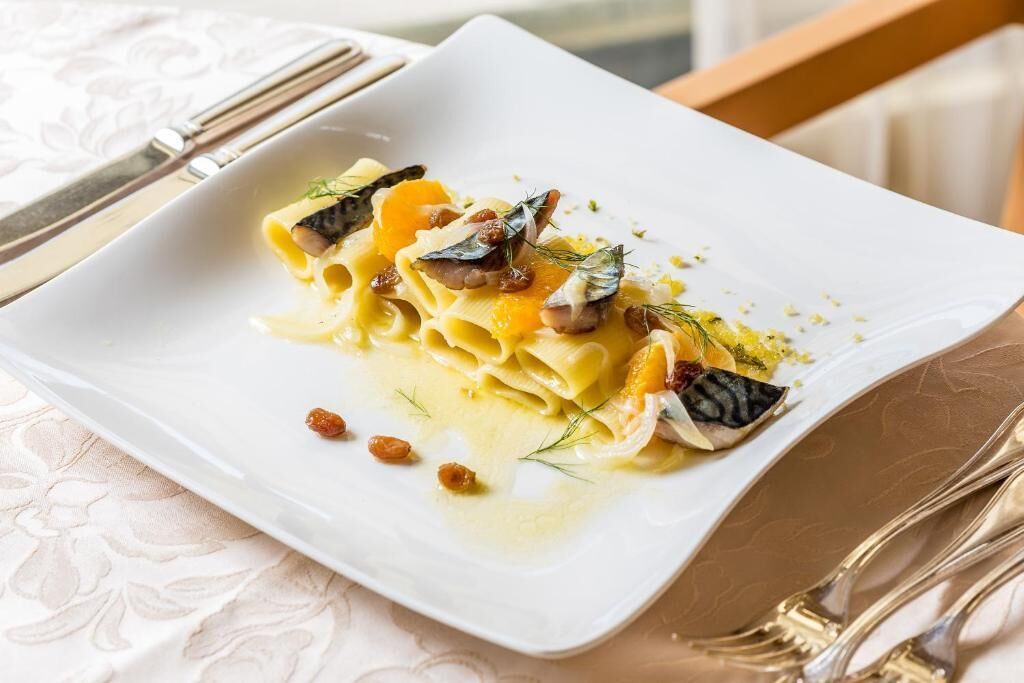 Une assiette carrée blanche contenant des pâtes, garnie d’herbes et de noix, posée sur une table recouverte d’une nappe blanche.