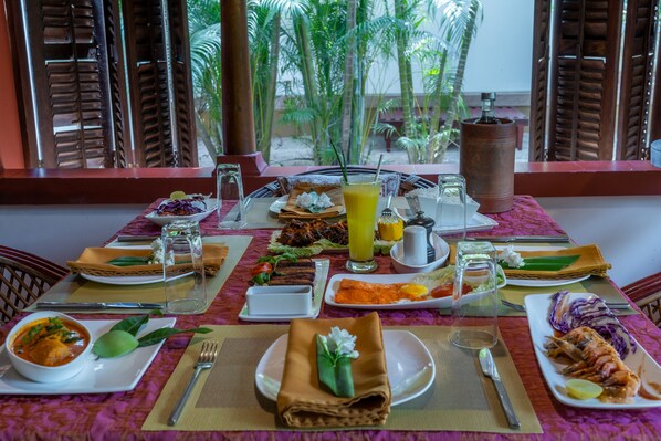 A table set with various dishes, a glass of orange juice, and a view of palm trees through wooden shutters. - Amritara A Beach Symphony (Cherthala)