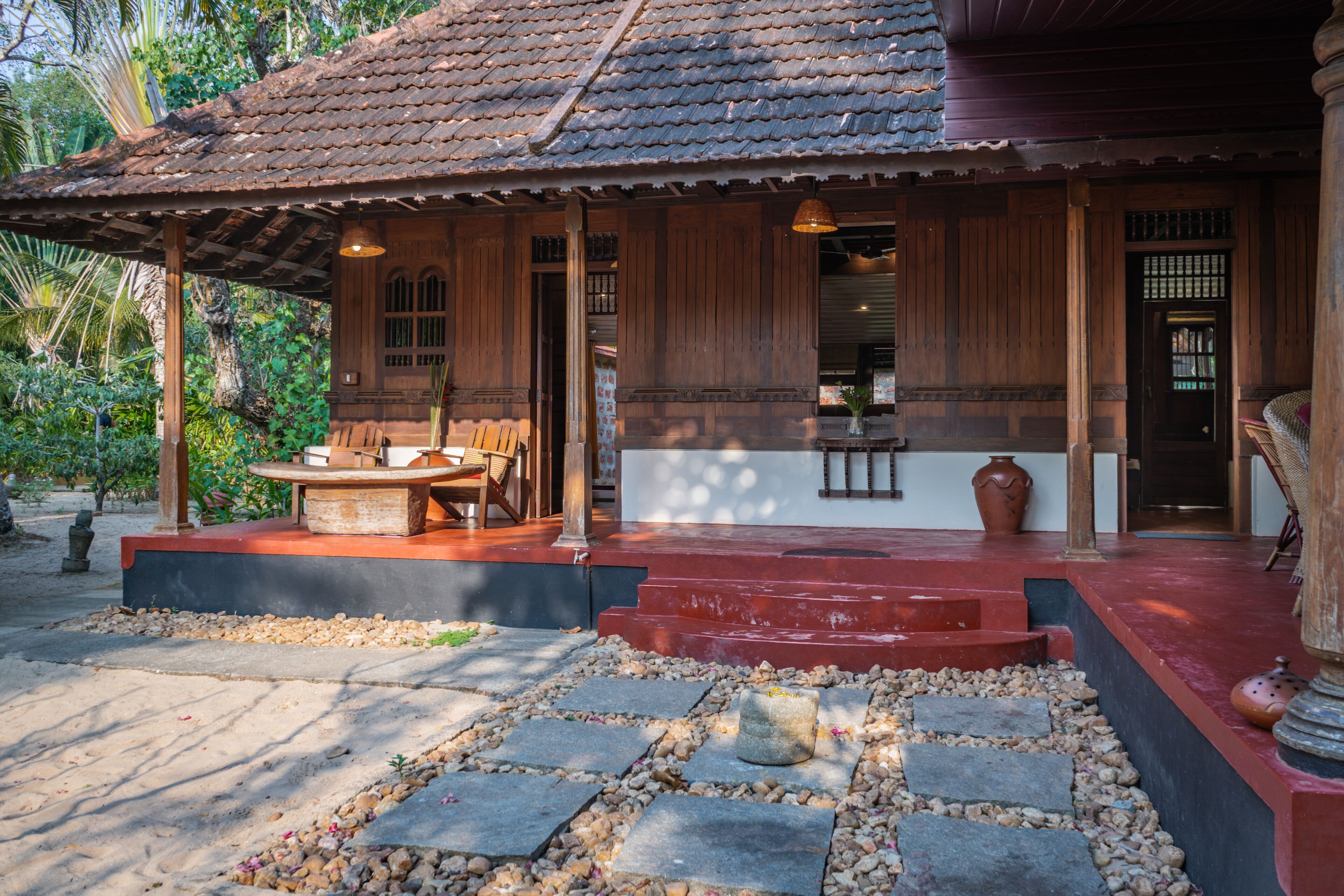A traditional wooden house with a tiled roof, a porch with a table and chairs, and a stone pathway leading to the entrance.