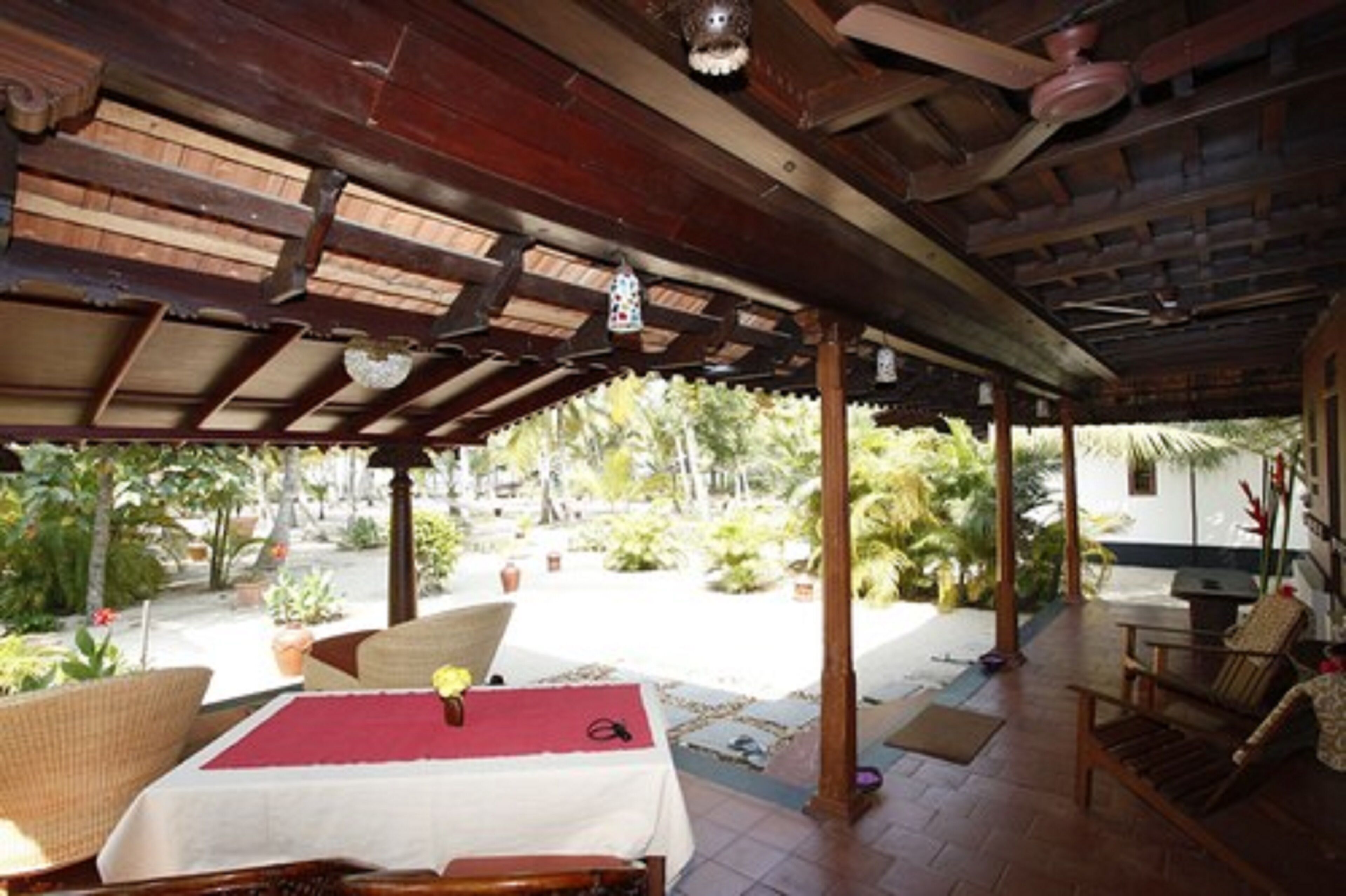 Uma área de estar coberta ao ar livre, com mesa e cadeiras, rodeada por plantas e vista para o exterior.