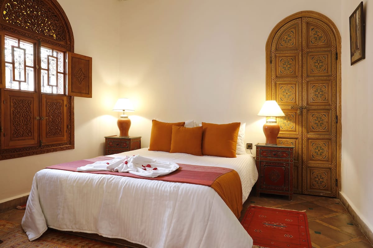 A neatly made bed with a white and red bedspread, two bedside lamps, and a large wooden door with intricate designs.