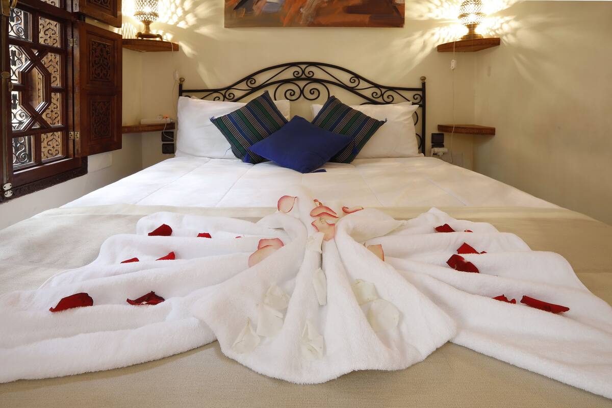 A neatly made bed with white linens and red rose petals, a wooden headboard, and two bedside lamps.
