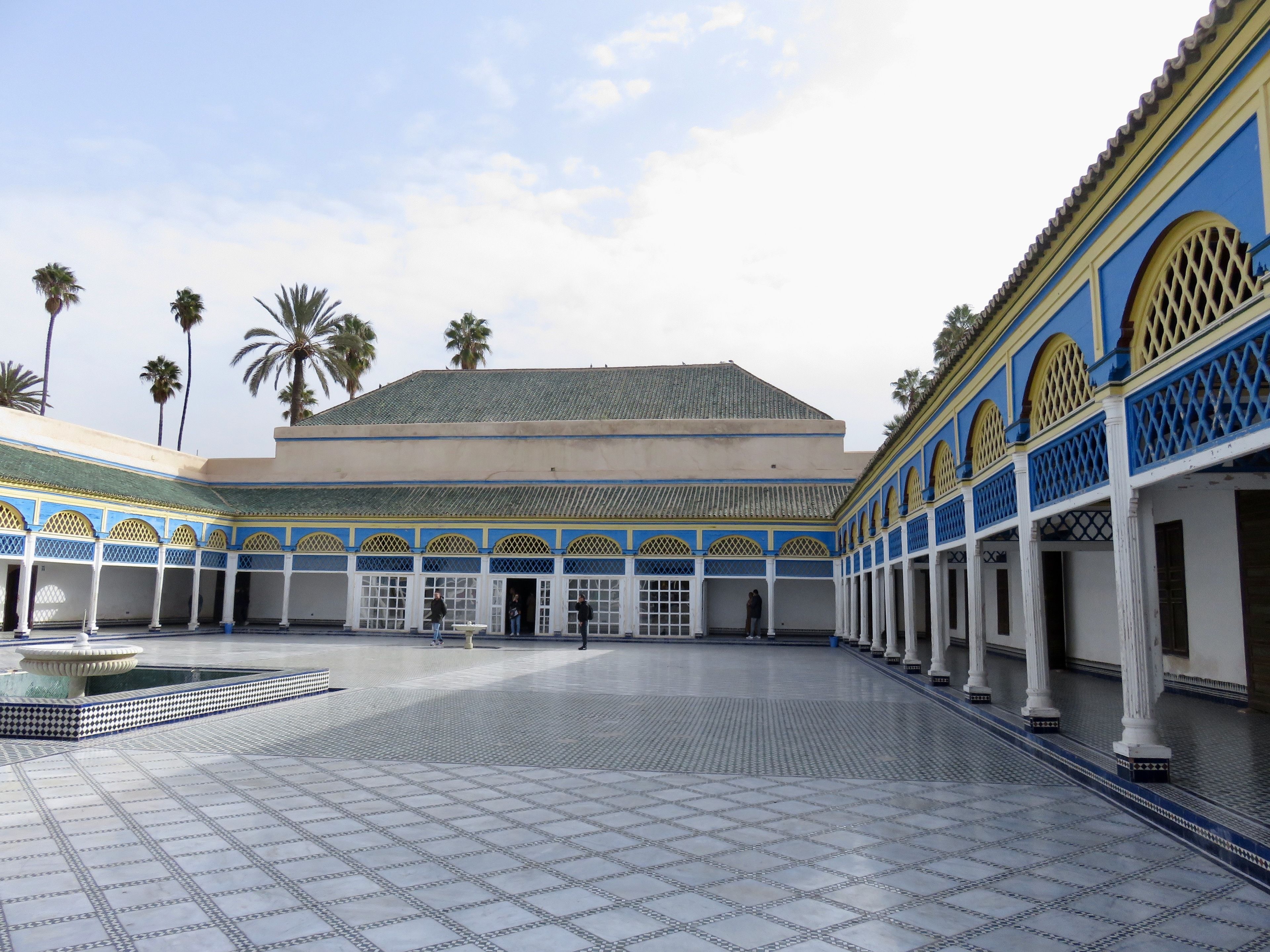 Un patio con suelo de baldosas, palmeras y un edificio con detalles arquitectónicos azules y amarillos.