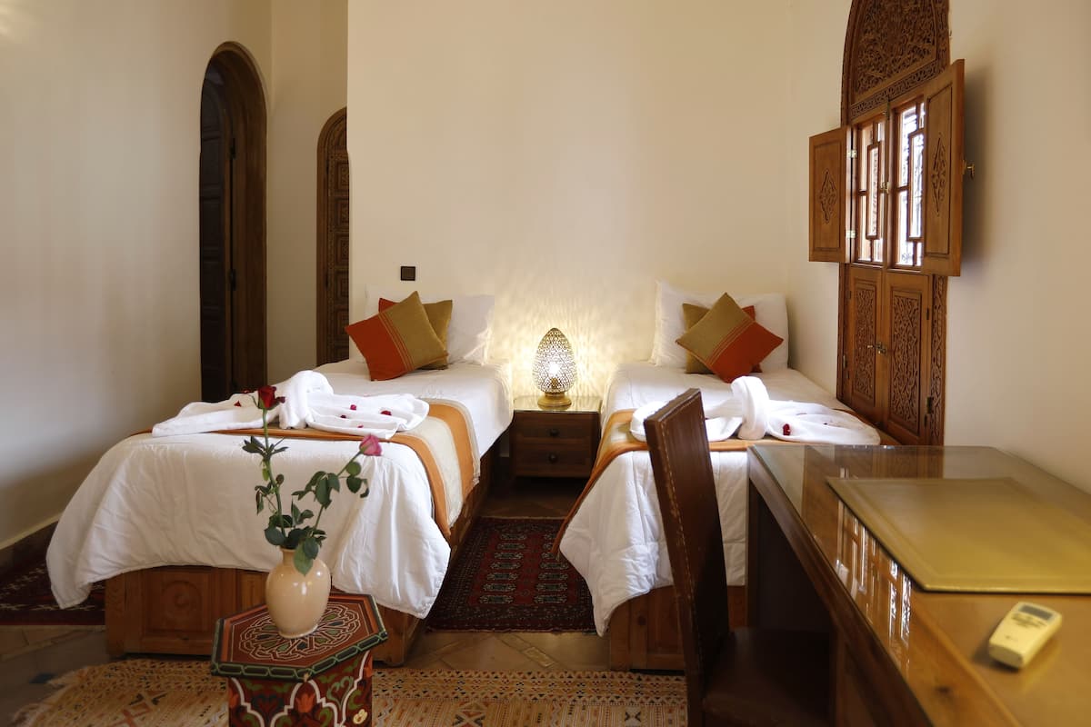 A hotel room with two beds, a small table, a chair, a decorative lamp, and a vase with a plant.