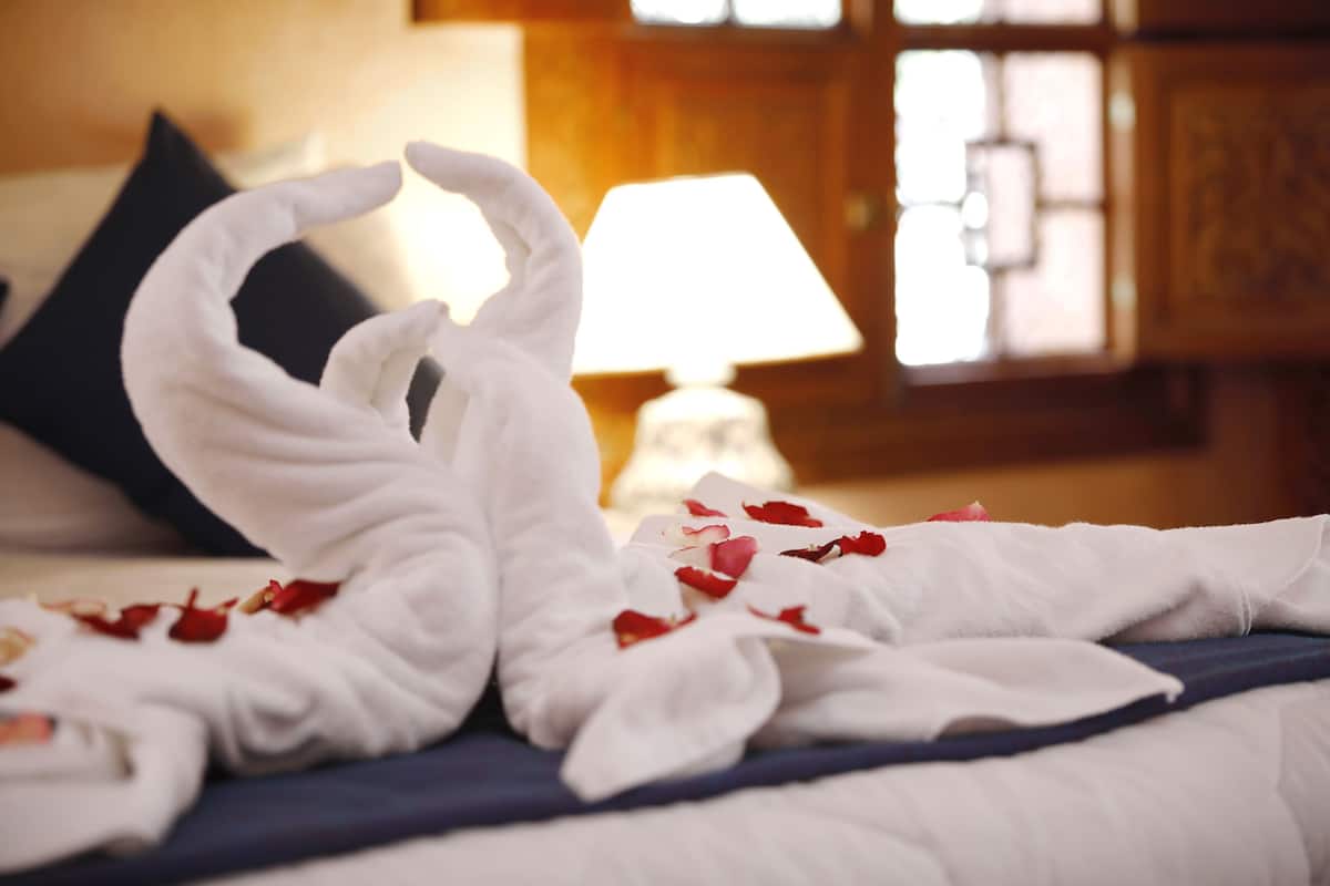 A bed with a white towel arranged in a swan shape, red rose petals scattered on it, and a dark blue bedspread.