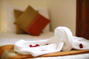 Una cama bien hecha con una toalla blanca y pétalos de rosa, un cabecero de madera y una almohada con estampado rojo y beige.