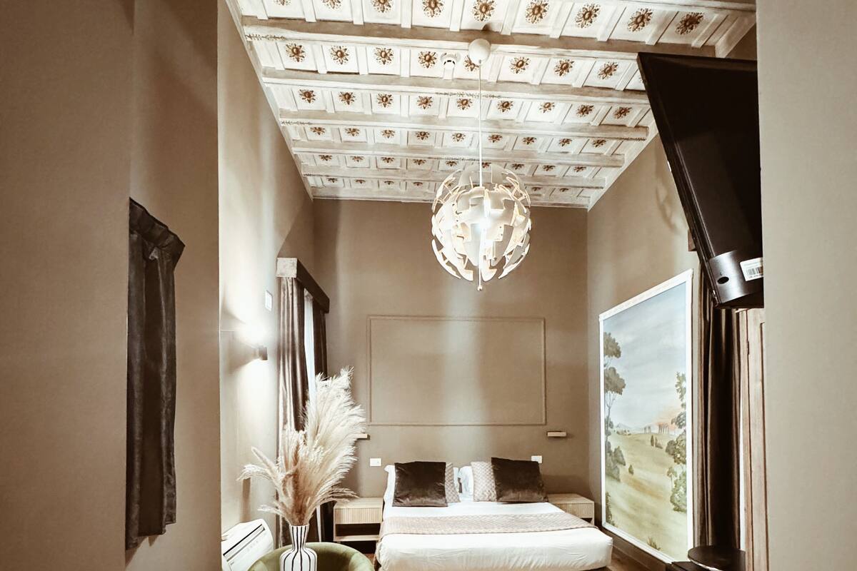 A bedroom with a wooden floor, a bed with white bedding, a green chair, a round table with a vase, a chandelier, and a painting on the wall.