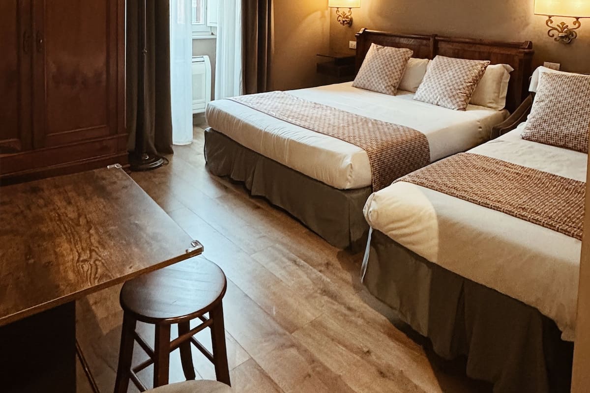 A hotel room with two beds, a wooden desk, a chair, a painting on the wall, and a view of the outside.