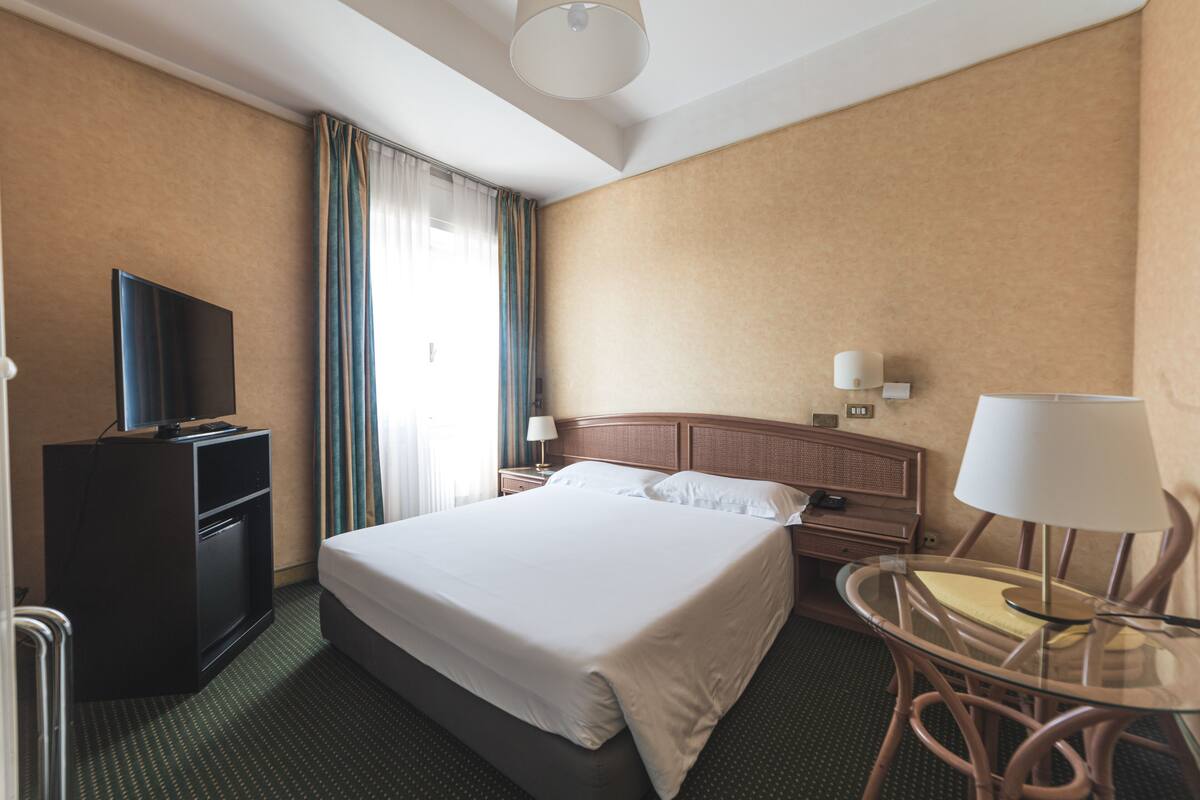 Ein Hotelzimmer mit einem großen Bett, einem Fernseher auf einem Schrank, einem Schreibtisch mit Lampe und einem Sessel.