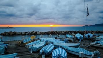 Una playa con sillones a rayas, barcos, un rompeolas rocoso y una puesta de sol sobre el agua.