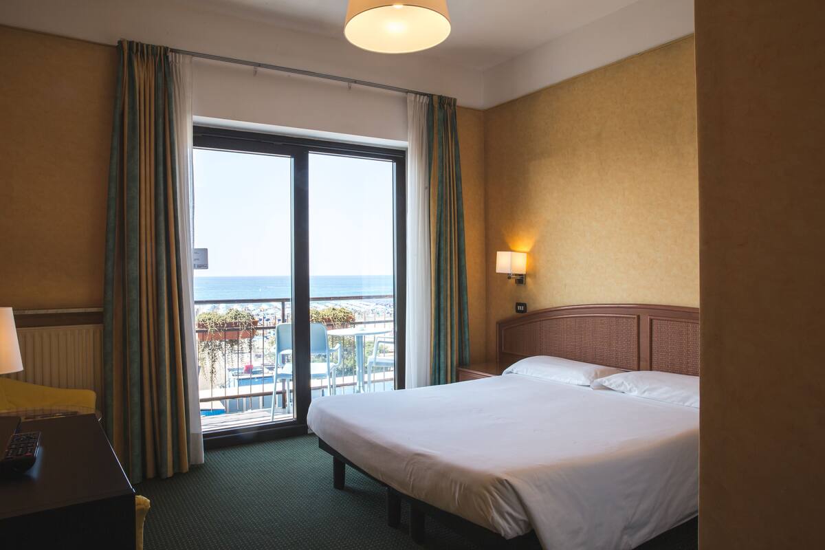 Ein Hotelzimmer mit Bett, Schreibtisch, Lampe, einem Fenster mit Vorhängen und Blick auf das Meer.