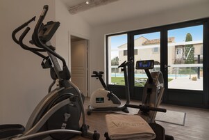 Un gimnasio en casa con equipo de ejercicio, un ventanal con vista a una casa y una piscina, y una puerta que da a otra habitación.