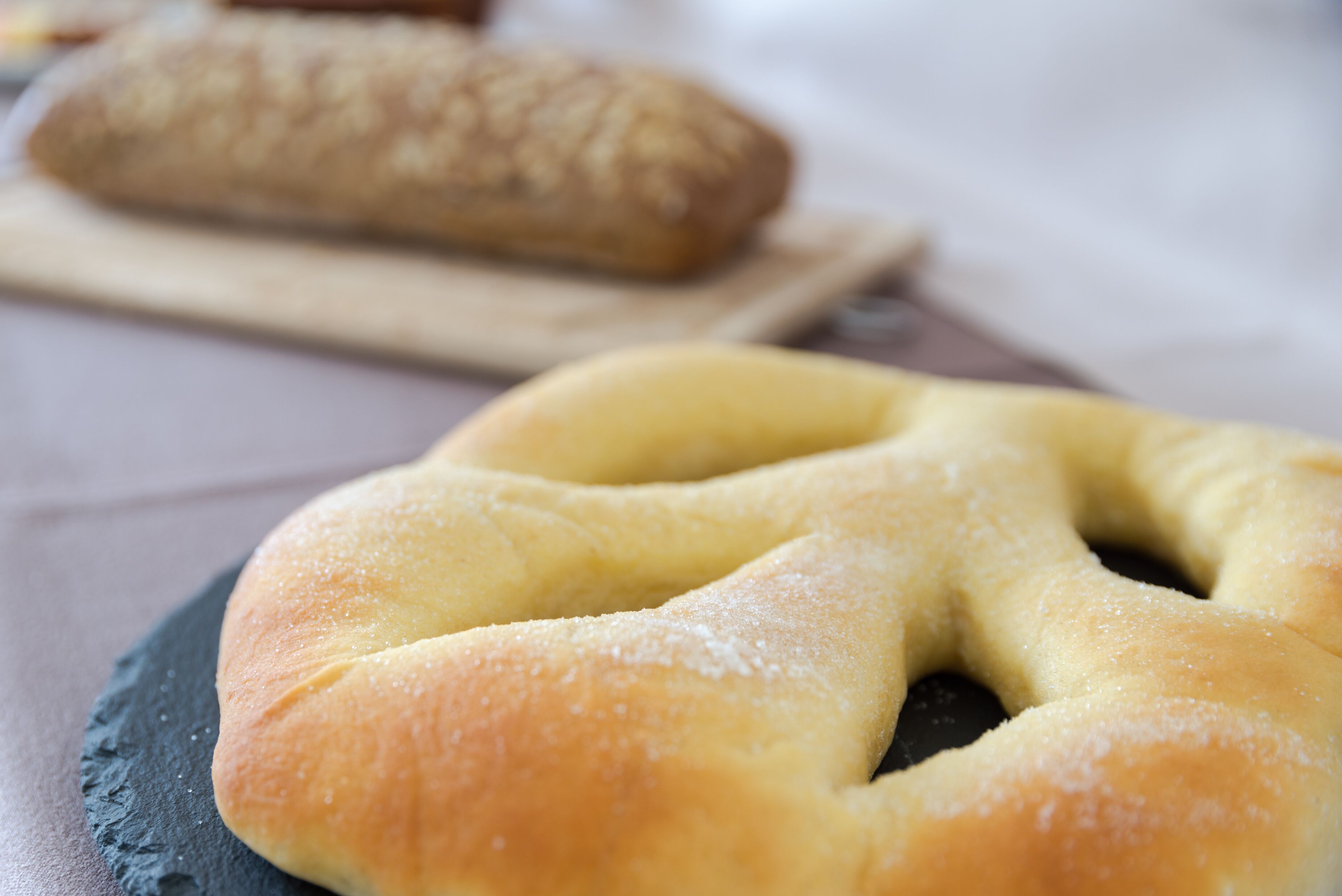 A close-up of a pretzel on a slate board with a loaf of bread in the background.